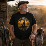 Papy portant un t-shirt noir avec le motif « PAPY CHASSEUR – Légende de la battue depuis toujours », photographié devant un chalet de chasse en forêt avec son chien, son équipement de chasse et un paysage boisé en arrière-plan.