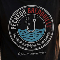Homme portant un t-shirt noir avec le design « Pêcheur Bredouille » affichant un air dépité au bord d'un lac après une journée de pêche sans prise.