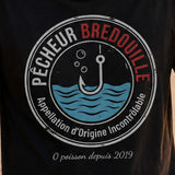 Homme portant un t-shirt noir avec le design « Pêcheur Bredouille » affichant un air dépité au bord d'un lac après une journée de pêche sans prise.