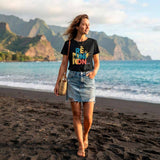 Une femme marche sur une plage de sable noir volcanique à La Réunion. Elle porte le t-shirt noir avec le design "RÉUNION" coloré et une jupe en jean claire. Elle a un sac en paille et des sandales. L'océan Indien est derrière elle, avec des montagnes luxuriantes au loin. La lumière est celle de la fin de l'après-midi.