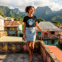 Une femme debout sur un toit-terrasse en béton, portant le t-shirt noir avec le design "LA REUNION LE LA" en blanc et bleu. Elle porte une jupe en jean claire et a les mains sur une rambarde métallique. L'arrière-plan montre des maisons colorées et des montagnes verdoyantes.