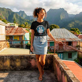 Une femme debout sur un toit-terrasse en béton, portant le t-shirt noir avec le design "LA REUNION LE LA" en blanc et bleu. Elle porte une jupe en jean claire et a les mains sur une rambarde métallique. L'arrière-plan montre des maisons colorées et des montagnes verdoyantes.