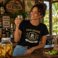 Femme portant un t-shirt noir avec le design « Rhum Arrangé » tenant un petit verre de rhum arrangé dans un bar créole à La Réunion