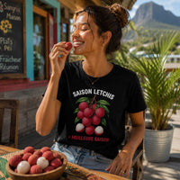 Femme portant un t-shirt noir avec le design « Saison Letchis = Meilleure Saison » dégustant un letchi sur la terrasse d'un café créole à La Réunion