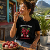 Femme portant un t-shirt noir avec le design « Saison Letchis = Meilleure Saison » dégustant un letchi sur la terrasse d'un café créole à La Réunion