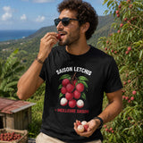 Homme portant un t-shirt noir avec le design « Saison Letchis = Meilleure Saison » en train de déguster un letchi dans un verger à La Réunion