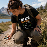 Femme portant un t-shirt noir avec l'inscription « Silence ça piste », examinant des empreintes d'animaux avec une loupe sur un sentier au bord d'un lac de montagne, entourée de sommets alpins et de fleurs sauvages.