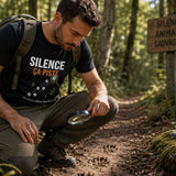 Homme portant un t-shirt noir avec l'inscription « Silence ça piste », concentré sur des empreintes d'animaux en forêt à l'aide d'une loupe, sur un sentier boisé au cœur de la nature.