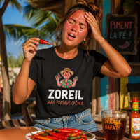 Femme portant un t-shirt noir avec le design « Zoreil mais presque créole », le visage rougi par la chaleur du soleil, tenant un piment rouge sur une terrasse tropicale.